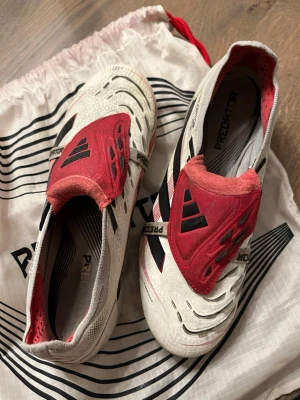 Adidas Predator vita och röda fotbollsskor - Säljer ett par Adidas Predator fotbollsskor i vitt och rött med svarta detaljer. Skorna har en modern design med täckt snörning och mönstrad ovandel för extra grepp på bollen. Perfekta för dig som vill dominera på planen.