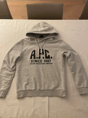 Grå hoodie från A.P.C. med tryck - Snygg ljusgrå hoodie från A.P.C. med svart tryck på bröstet där det står 'A.P.C. SINCE 1987'. Klassisk passform med huva och dragsko. Tillverkad i mjuk bomullsblandning, perfekt för en chill och stilren look.