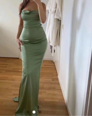 Grön långklänning med slits och snörning - Snygg grön långklänning i satinliknande material med smala axelband, draperad urringning och hög slits på sidan. Klänningen har öppen rygg med snörning och figurnära passform som framhäver siluetten. Klänningen har defekter som syns på de sista bilderna o är lite noppig . Köpt för 1100kr