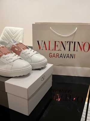 Valentino Rockstud sneakers vit/rosa - Snygga Valentino Rockstud sneakers i vitt skinn med rosa band och ikoniska nitar. Klassisk rund tå, platt sula och vita snören. Märkeslogga på plösen och cool design som sticker ut. Perfekta för dig som vill ha något extra till din outfit.