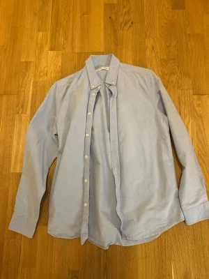 Ljusblå skjorta från Samsøe Samsøe - Klassisk ljusblå skjorta från Samsøe Samsøe i mjukt bomullstyg. Skjortan har knappar framtill, lång ärm och en stilren krage. Perfekt för en clean och avslappnad look.