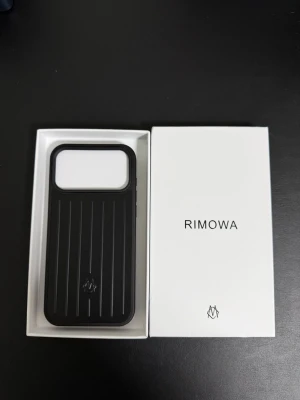 Svart Iphone 17 pro max Rimowa mobilskal  - Snyggt svart mobilskal från Rimowa med klassisk räfflad design och diskret logotyp. Skalet är tillverkat i tåligt material och har en stilren, modern look som passar perfekt till din iPhone.
