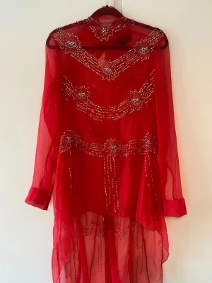 Röd långklänning med paljettdetaljer - Snygg röd asymmetrisk klänning/tunika i transparent material med långa ärmar och glittriga paljettdetaljer.  Perfekt layering piece! 