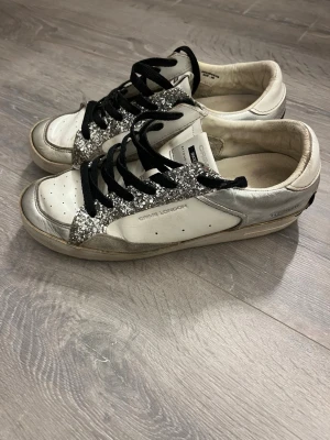 Glittriga sneakers från Crime London - Snygga sneakers från Crime London i vitt och silver med glittriga detaljer och svarta skosnören. Skorna har rund tå och platt sula, samt en cool kontrast med svart hälparti. Perfekta för dig som vill sticka ut med lite extra bling.