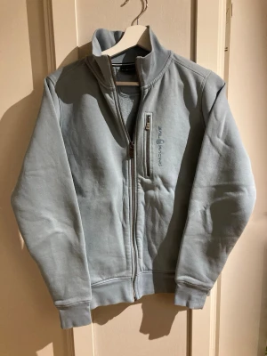 Ljusgrå ziptröja från Sail Racing - Ljusgrå ziptröja från Sail Racing med hög krage och broderad logga på bröstet. Tröjan har hel dragkedja framtill, ribbade muddar och en sportig look. Perfekt för lager-på-lager och chill vardagsstil.