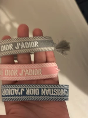 Christian Dior J'Adior tygarmband - Säljer tre snygga tygarmband från Christian Dior med texten 'J'ADIOR'. Armbanden kommer i grått, rosa och blått med vita detaljer och har en trendig tofs i änden. Perfekta för att ge din outfit en exklusiv touch.