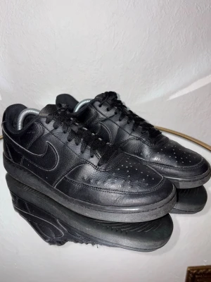 Svarta Nike Air Force 1 sneakers - Klassiska helsvarta Nike Air Force 1 sneakers i skinn med rund tå och snörning. Swoosh-logga på sidan och perforerade detaljer på tån för extra ventilation. Tidlös och clean design som passar till det mesta.