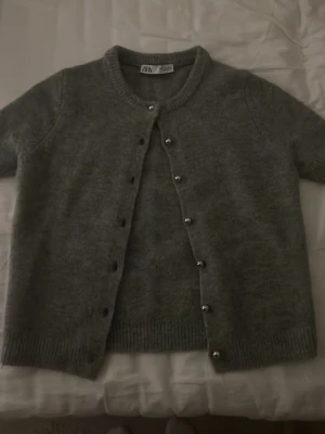 Grå kofta från Zara med knappar - Mysig grå kofta från Zara med rund halsringning och korta ärmar. Koftan har ribbade muddar och snygga knappar framtill. Perfekt att slänga på sig över en t-shirt eller linne. säljer då dem ej används. 