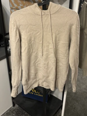 Soft Goat hoodie - Beige stickad hoodie från Soft Goat i mjuk och lyxig kvalitet. Tröjan har huva med dragsko och ribbstickade muddar vid ärmslut och nederkant. Perfekt för en avslappnad och stilren look. Nypris 3500k:-