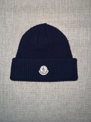 Marinblå ribbstickad Moncler mössa - Säljer en marinblå ribbstickad mössa från Moncler! Perfekt för lite kyligare dagar då den är gjord i ull och inte är kall men du blir inte för varm! Mössan är helt oanvänd och riktigt bra skick! Hör av er vid intresse, snabb leverans!!