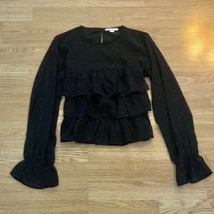 Svart volangblus  - Svart glittrig blus från Gina Tricot. Långärmad modell med lätt utsvängda ärmslut och rund halsringning.