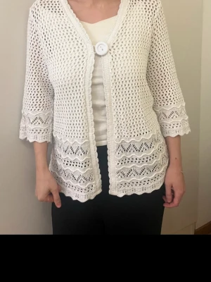 Vit virkad kofta med knapp - Snygg vit stickad kofta med virkat mönster och trekvartsärmar. Koftan har en stor dekorativ knapp framtill och vågiga kanter nertill och vid ärmslut. 