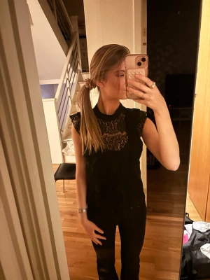 Svart spetsblus med volangdetalj - Säljer en svart blus i spets med hög hals och volangdetaljer på axlarna. Blusen är ärmlös och har ett elegant, transparent mönster upptill. Perfekt för dig som gillar en stilren och trendig look.