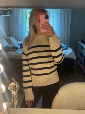 Beige stickad tröja med svarta ränder - Snygg stickad tröja i beige med breda svarta ränder. Tröjan har en högre ribbad krage, långa ärmar och ribbade muddar vid ärmslut och nederkant. Perfekt för en trendig och avslappnad look.