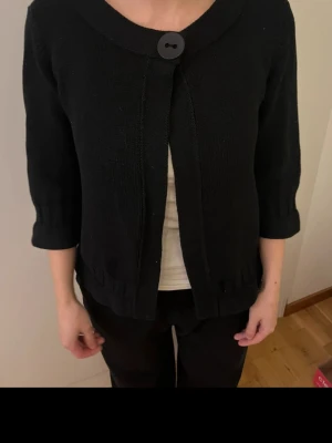 Svart stickad kofta  - Svart stickad kofta med  trekvartsärm och stor dekorativ knapp framtill. Jackan har en rundad krage och en rak, något loose passform. 