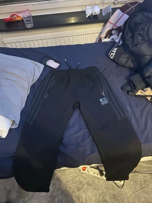 Svarta adidas byxor - Svarta mjukisbyxor från Dope med dragsko i midjan och två dragkedjeförsedda fickor framtill. Byxorna har en diskret logga på vänster ben och är tillverkade i ett mjukt material som passar perfekt för chill eller träning. Adidas byxor 