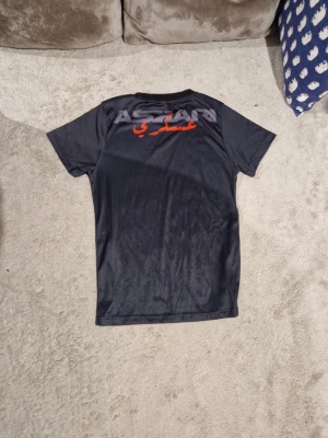 Svart t-shirt med tryck från Askar - Svart t-shirt med rund hals och tryck i vitt och rött både fram och bak. Tillverkad i ett glansigt, mjukt material som känns sportigt och bekvämt. Perfekt för dig som gillar stilrena plagg med en twist.