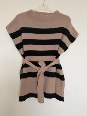 Randig beige och svart stickad väst - Stickad väst i beige och svart med breda horisontella ränder och knytskärp i midjan. Modellen är ärmlös med rund halsringning och har en avslappnad, boxig passform. Perfekt att styla över en långärmad tröja för en trendig lager-på-lager-look.