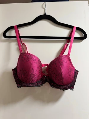 Rosa push-up bh med spetsdetaljer - Snygg rosa push-up bh med svarta spetsdetaljer och blommigt mönster. Bh:n har vadderade kupor, justerbara axelband och en liten rosett med pärlberlock framtill. Materialet är glansigt och ger en lyxig känsla.