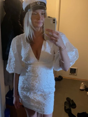 Vit kort klänning med puffärmar - Superfin vit kort klänning med djup v-ringning och stora puffärmar. Klänningen har en strukturerad yta och markerad midja som ger en snygg siluett. Perfekt för student eller sommarens alla tillfällen.