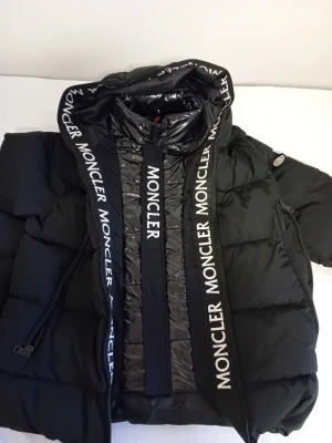 Svart dunjacka från Moncler - Snygg svart dunjacka från Moncler med stor huva och tydliga logoband längs dragkedjan. Jackan har två snedställda fickor med dragkedja och quiltad design. Perfekt för kalla dagar och ger en riktigt trendig look. Jackan har används en vinter och har hängt I garderoben I 3 år. Jackan är storlek M/L och har bra passform.