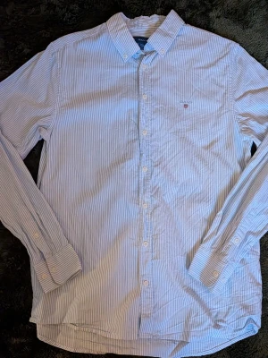 Blårandig skjorta från Gant stl 170 cm  - Klassisk blårandig skjorta från Gant med vit knappstängning och liten broderad logga på bröstet. Skjortan har lång ärm, krage och är tillverkad i mjuk bomull. Perfekt för en clean och stilren look.