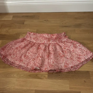 Rosa mönstrad kortkjol - Säljer en luftig kortkjol med rödvitt paisleymönster. Kjolen har en bred midjeresår och är gjord i ett tunt, skönt bomullstyg som passar perfekt till varma dagar. Snygg att matcha med en enkel topp eller hoodie.