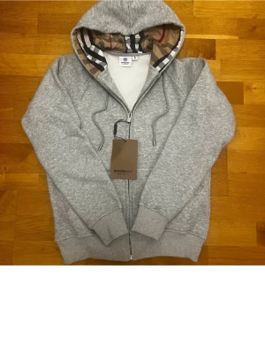 Grå hoodie från Burberry - Grå zip-hoodie från Burberry med klassiskt rutigt foder i huvan och dragsko. Mjuk bomullsblandning och ribbade muddar. Perfekt för en chill och stilren look.