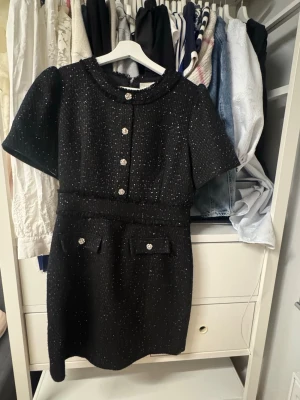 Svart kortklänning med glittriga knappar - Stilren svart kortklänning med glittriga knappar framtill och dekorativa fickor. Klänningen har korta puffärmar och en rund krage. Materialet är strukturerat och ger en exklusiv känsla.