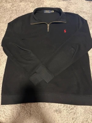Svart halvzip från Polo Ralph Lauren - Svart långärmad tröja från Polo Ralph Lauren med halv dragkedja och röd broderad logga på bröstet. Klassisk ribbad mudd vid ärmslut och nederkant. Perfekt för en clean och stilren look.
