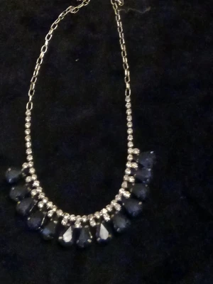 Silverfärgat halsband med blå droppar - Snyggt halsband med silverfärgad kedja och glittrande stenar. Framhävs av stora blå droppformade hängen och små klara stenar längs hela framsidan. Perfekt för att ge din outfit lite extra bling. Materialet är troligen metall och glas.