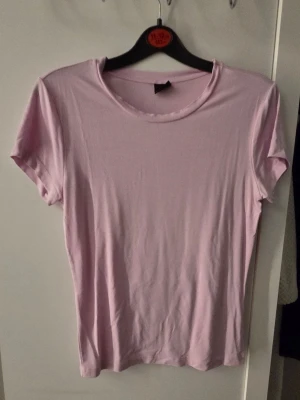 Ljusrosa basic t-shirt från Gina Tricot  - En enkel och stilren ljusrosa t-shirt med rund hals och korta ärmar. Perfekt att matcha med jeans eller kjol för en clean look. Tillverkad i mjukt material som känns skönt mot huden.