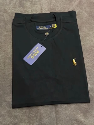 Svart t-shirt från Polo Ralph Lauren - Klassisk svart t-shirt från Polo Ralph Lauren med gul broderad logga på bröstet. T-shirten har rund halsringning och korta ärmar. Tillverkad i mjuk bomull för en skön känsla och enkel stil.