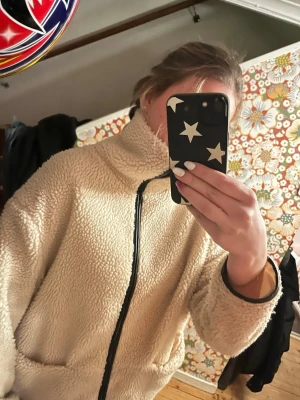 Beige teddyjacka med dragkedja - Mysig beige teddyjacka med hög krage och svarta detaljer längs dragkedjan och ärmsluten. Jackan har två stora fickor framtill och en avslappnad, lite oversized passform. Perfekt för kyliga dagar när du vill vara både varm och snygg.