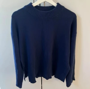 Mörkblå stickad tröja - Mörkblå stickad tröja med bred ribbad kant vid hals, ärmslut och nederkant. Tröjan har en avslappnad passform och långa ärmar, perfekt för kyliga dagar. Enkel och stilren design som funkar till det mesta.