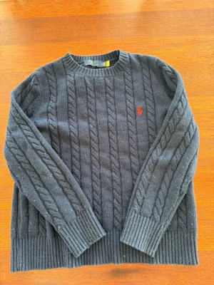 Mörkblå kabelstickad tröja Polo Ralph Lauren - Kabelstickad mörkblå tröja från Polo Ralph Lauren med rund halsringning och ribbade muddar. Tröjan har det klassiska röda Polo-logot broderat på bröstet och är tillverkad i mjukt material. Perfekt för dig som gillar tidlös och stilren design.