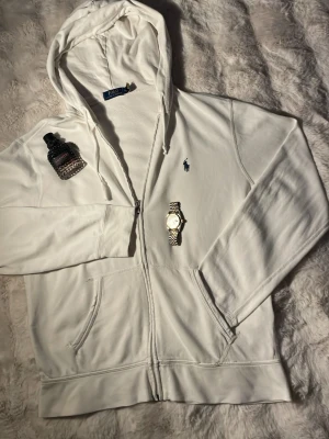 Vit hoodie från Polo Ralph Lauren - Snygg vit hoodie från Polo Ralph Lauren med dragkedja och klassisk blå logga på bröstet. Hoodien har huva med dragsko och två fickor framtill. Tillverkad i mjukt bomullsmaterial som känns skönt mot huden. Perfekt för en clean och stilren look. Storlek M så skulle rekommendera dig som är runt 170-180, nypris runt 1500 KR