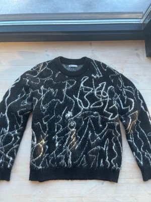 Svart stickad tröja med mönster från Zara strl L - Cool svart stickad tröja från Zara med vitt abstrakt mönster över hela plagget. Tröjan har rund halsringning och ribbade muddar vid ärmslut och nederkant. Materialet är fluffigt och ger en trendig look.