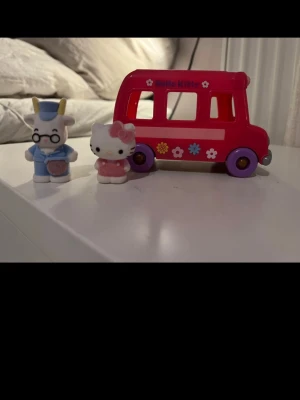 Hello Kitty leksaksbuss med figurer - Säljer en vintage röd Hello Kitty leksaksbuss i plast med blommor på sidorna och två små figurer, en Hello Kitty och en blåklädd kanin. Perfekt för samlare eller som rolig leksak till barnrummet.