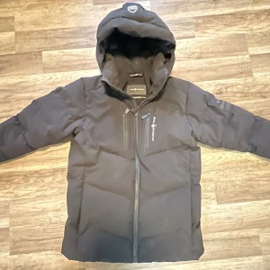 Svart pufferjacka från sailracing - Svart pufferjacka i nytt skick från sailracing med huva och dragkedja framtill. Jackan har en bröstficka med dragkedja och diskret logga på bröstet. Perfekt för kalla dagar med sin vadderade design och långa ärmar. 