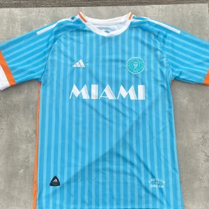 Inter Miami Messi 10 Adidas fotbollströja - Säljer en blå och orange Inter Miami fotbollströja från Adidas med Messi 10 på ryggen. Tröjan har vita detaljer, ränder och klubbmärke på bröstet samt tryck med MIAMI på framsidan. Korta ärmar och tillverkad i lätt, ventilerande material.