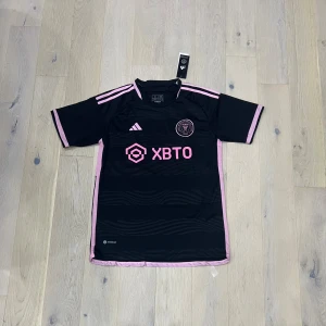 Inter Miami svart och rosa matchtröja - Snygg Inter Miami fotbollströja från Adidas i svart med rosa detaljer och logga. Kortärmad modell med rosa ränder på axlarna och rosa kant vid ärmar och nederkant. Tillverkad i lätt och ventilerande polyester, perfekt för träning eller match.