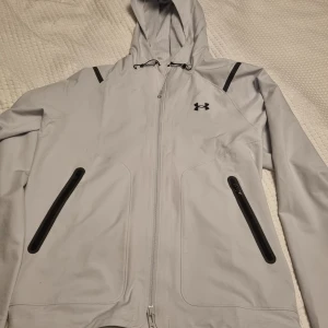 Ljusgrå jacka Under Armour - Ljusgrå vindjacka från Under Armour med huva och svarta detaljer. Jackan har dragkedja framtill och två snedställda fickor. Se gärna mina andra annonser och skapa bundles. Inga byten. 