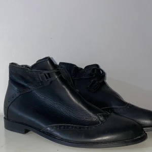 Svarta skinnboots med spänne, stl 38 - Snygga svarta boots i skinn med dekorativa broguedetaljer och spänne framtill. Skorna har låg klack, rund tå och dragkedja på sidan för enkel påtagning. Perfekta för dig som gillar klassisk stil med en twist.
