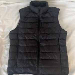 Svart dunväst från Uniqlo Ultra Light Down - Svart, lätt och quiltad dunväst från Uniqlo Ultra Light Down. Vill bara sälja den, därför billigt. Västen har hög krage, dragkedja framtill och medföljande förvaringspåse. Perfekt för lager-på-lager och enkel att packa ner i väskan.