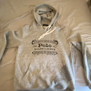 Grå hoodie från Polo Ralph Lauren - Snygg grå hoodie från Polo Ralph Lauren med svart tryck framtill. Klassisk känguruficka, vit snörning i huvan och mjukt material. Perfekt för chill dagar och streetstyle. Regular fit och långärmad modell.