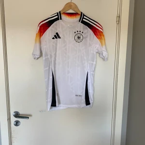 Tysklands landslagströja Adidas M - Säljer en officiell Tyskland fotbollströja från Adidas i storlek M. Tröjan är vit med svarta, röda och gula detaljer på axlarna, har korta ärmar och ett diskret geometriskt mönster. Märkt med HEAT.RDY-teknologi för extra komfort. Slim fit-modell.