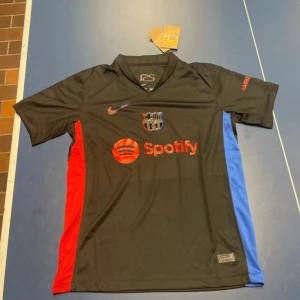 Svart FC Barcelona Nike fotbollströja - Snygg svart FC Barcelona fotbollströja från Nike med röda och blå detaljer på sidorna. Tröjan har klubbmärke och Nike-logga med röd och blå kant, samt Spotify som sponsor på bröstet.