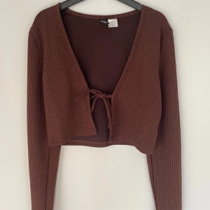 Brun ribbad bolerotopp från H&M Divided - Säljer en brun ribbad bolerotopp från H&M Divided i storlek XS. Toppen är croppad med långa ärmar, v-ringning och knyt framtill. Perfekt för att slänga över ett linne eller klänning för en trendig lager-på-lager-look.