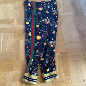 Gucci mönstrade mjukisbyxor med ränder - Gucci mjukisbyxor i mörkblått med färgglatt mönster av solar, planeter och fåglar. Sidorna har gröna och röda ränder, och bensluten är ribbade i gult med blå ränder. Byxorna har en avslappnad passform och är tillverkade i mjukt material.